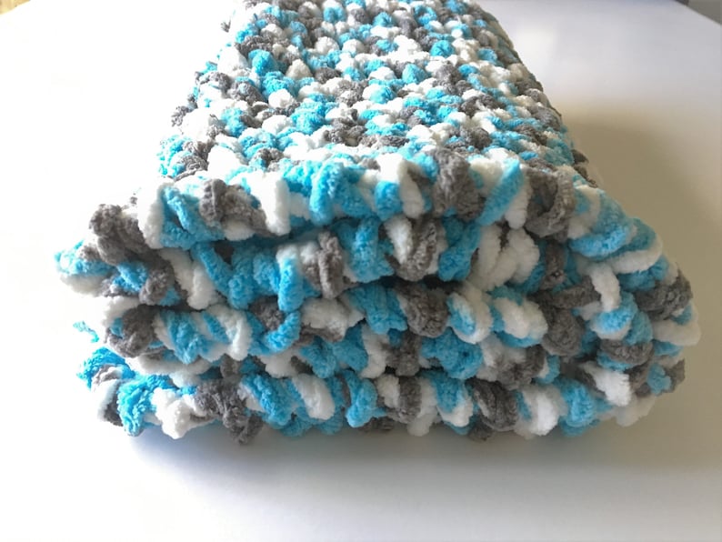 May include: A folded crocheted baby blanket in shades of blue, white, and gray. The blanket is made with a soft, fluffy yarn.