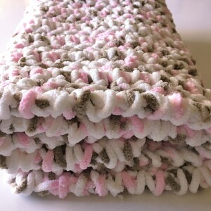 Puede incluir: Una suave manta de beb&eacute; tejida a crochet en rosa, blanco y marr&oacute;n. La manta est&aacute; doblada en tres partes y est&aacute; hecha con un hilo grueso.