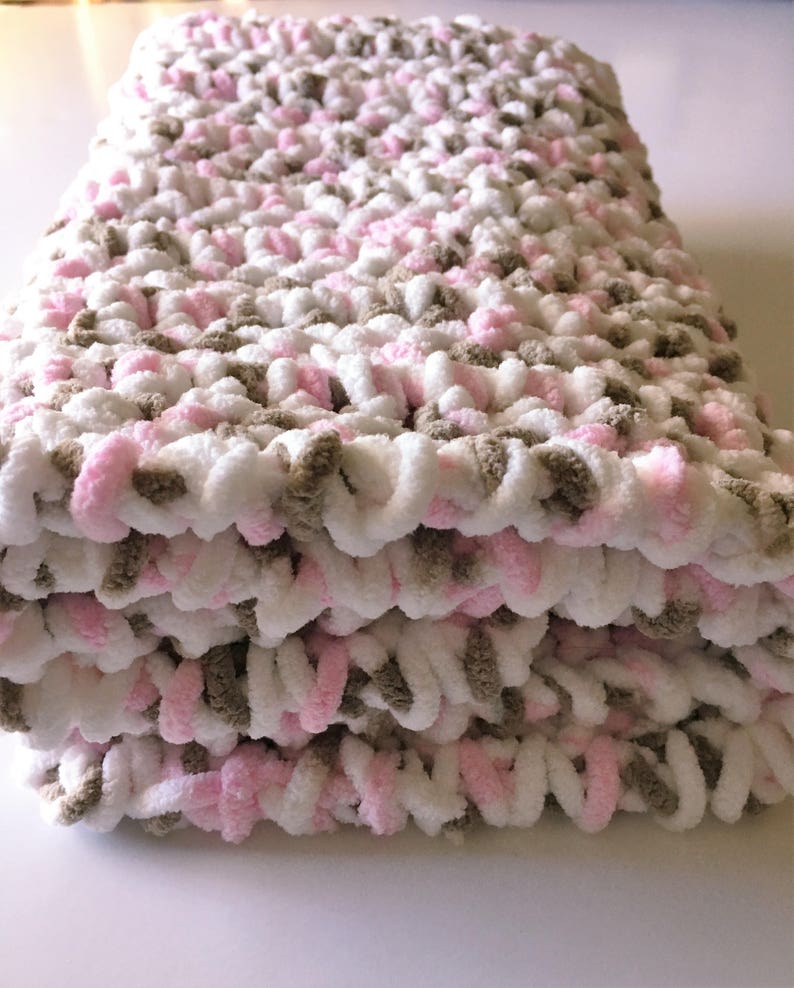 Puede incluir: Una suave manta de beb&eacute; tejida a crochet en rosa, blanco y marr&oacute;n. La manta est&aacute; doblada en tres partes y est&aacute; hecha con un hilo grueso.