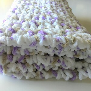 May include: A soft, crocheted baby blanket in white, lavender, and beige. The blanket is folded in half and has a fluffy texture.