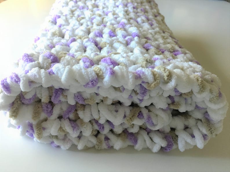 May include: A soft, crocheted baby blanket in white, lavender, and beige. The blanket is folded in half and has a fluffy texture.