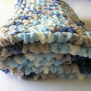 May include: A soft, crocheted baby blanket in shades of blue, white, and gray. The blanket is folded and has a fluffy texture.