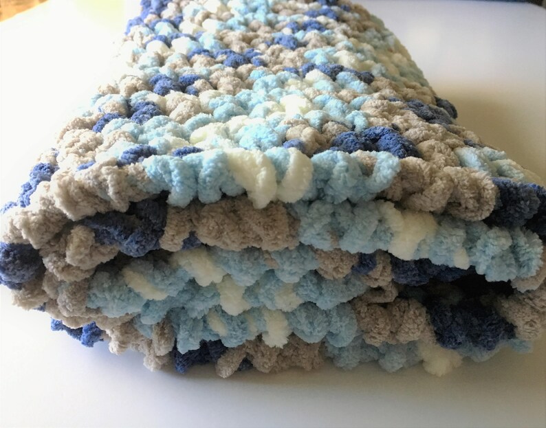 May include: A soft, crocheted baby blanket in shades of blue, white, and gray. The blanket is folded and has a fluffy texture.