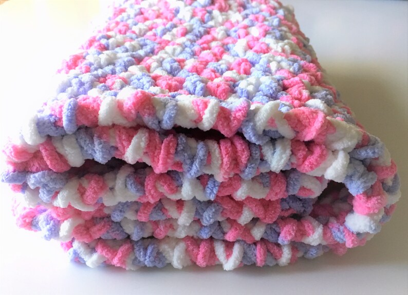 Puede incluir: Una manta de beb&eacute; de crochet rosa, blanca y lavanda doblada. La manta est&aacute; hecha con un hilo suave y esponjoso.