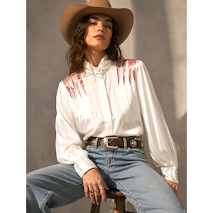 May include: A tan cowboy hat, a white long-sleeved button-down shirt with pink satin shoulder accents, and light blue denim jeans with a brown leather belt featuring silver concho hardware. The shirt has a high collar and long cuffed sleeves.