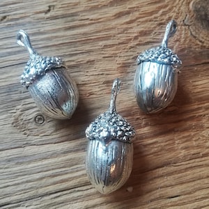 Könnte beinhalten: Drei silberne Eichel-Anhänger mit detaillierten Texturen. Jeder Anhänger hat eine Öse zum Anbringen an einer Halskette, einem Armband oder anderem Schmuck.