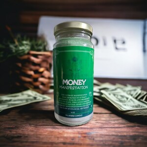 May include: Green glass jar candle with a gold lid. The label reads "NATURALLY YOU MONEY MANIFESTATION The ultimate manifestation tool designed to attract abundance, wealth and prosperity to your life. This candle harnesses the power of natural herbs and crystals to amplify your intentions and unlock the flow of financial abundance. 16 oz PALMWAX CANDLE"