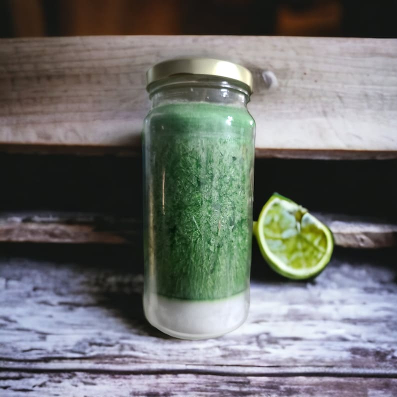 May include: A clear glass jar with a gold lid filled with a green candle. The candle has a white layer at the bottom. A half of a lime is sitting on a wooden surface next to the jar.