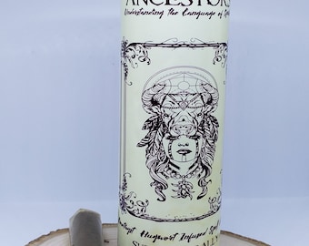 May include: Purple candle with a white label that reads "ANCESTORS Understanding the Language of Spirits", a black and white illustration of a woman with a bull's head, and the text "Highest Hugwort Infused Spell SUPERNATURALLY YOU". The candle is sitting on a wooden surface with a brown cigar.
