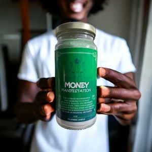 May include: A green and gold candle with the words "MONEY MANIFESTATION" on the label. The candle is designed to attract wealth and prosperity. It is a palm wax candle and is 16 oz.
