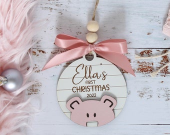 May include: A pink and white wooden ornament with the text "Ella's First Christmas 2022" and a pink pig illustration.