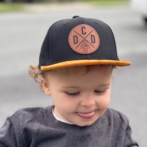 Peut inclure: Une casquette de baseball noire et marron avec un patch en cuir marron représentant un robot et les lettres "DCD" dans un cercle. La casquette a une visière marron.