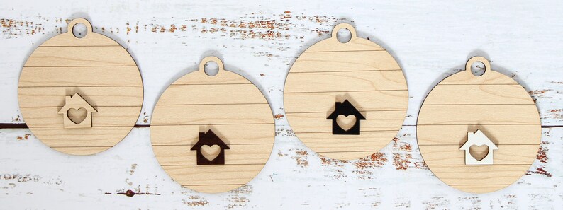 May include: Four wooden ornaments in the shape of a circle with a house design in the center. The houses have a heart in the center and are in different colors: white, black, and brown. The ornaments are perfect for decorating a home for the holidays.