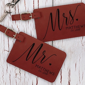 K&ouml;nnte beinhalten: Zwei rote Leder-Gep&auml;ckanh&auml;nger mit schwarzer Beschriftung. Der obere Anh&auml;nger lautet "Mrs. MATTHEWS Est. 2019" und der untere Anh&auml;nger lautet "Mr. MATTHEWS Est. 2019".
