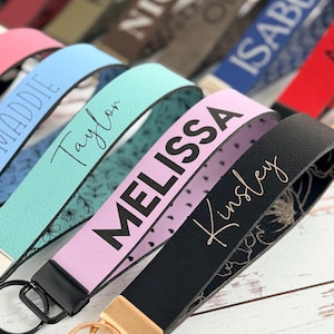 Assorted personalized leatherette wristlet keychains with engraved names (Taylor, Melissa, Kinsley) in blue, teal, lavender, and black, shown close-up with metal key rings on a white wood surface; more name straps blurred behind.