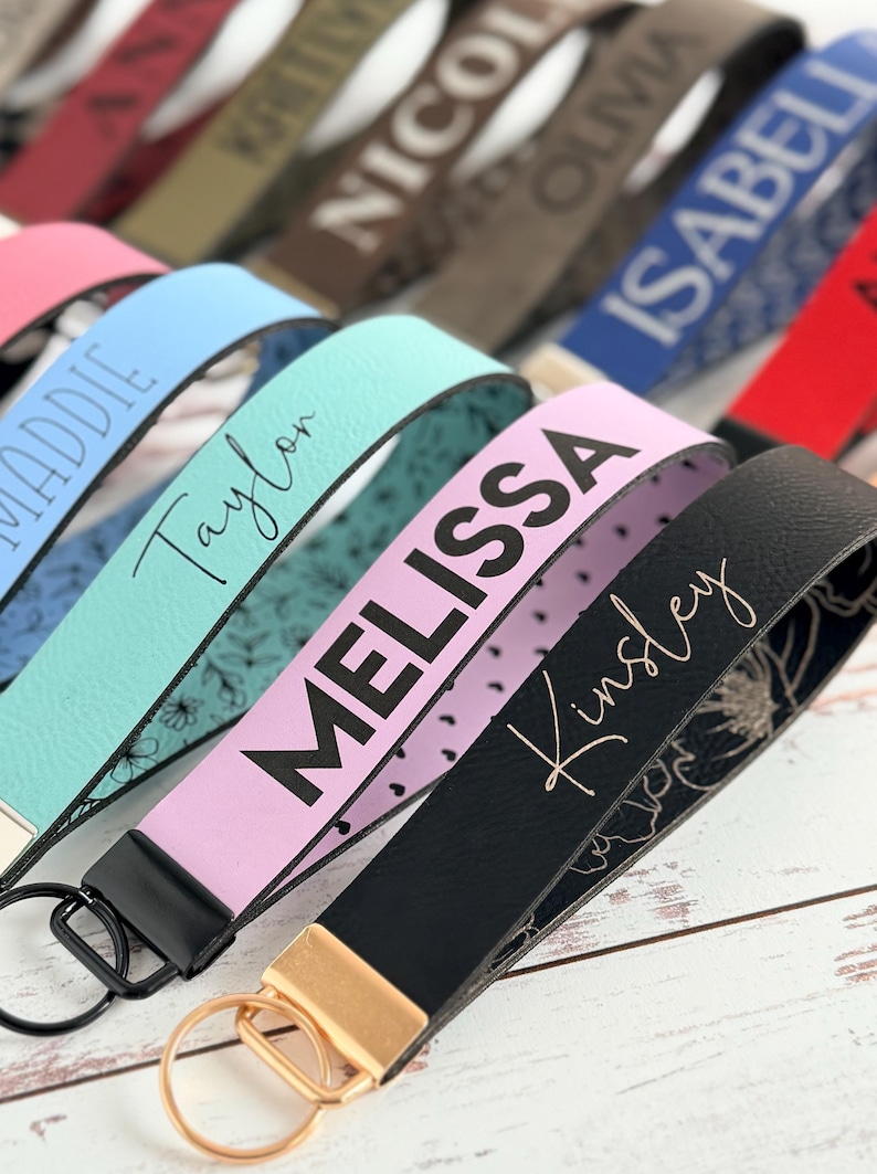 Assorted personalized leatherette wristlet keychains with engraved names (Taylor, Melissa, Kinsley) in blue, teal, lavender, and black, shown close-up with metal key rings on a white wood surface; more name straps blurred behind.