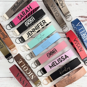 May include: A collection of personalized keychains in various colors, including pink, black, and blue. Each keychain features a name in a contrasting color, such as "Sarah" or "Jennifer", and includes a metal ring for attaching keys. The keychains are arranged on a light wood surface.