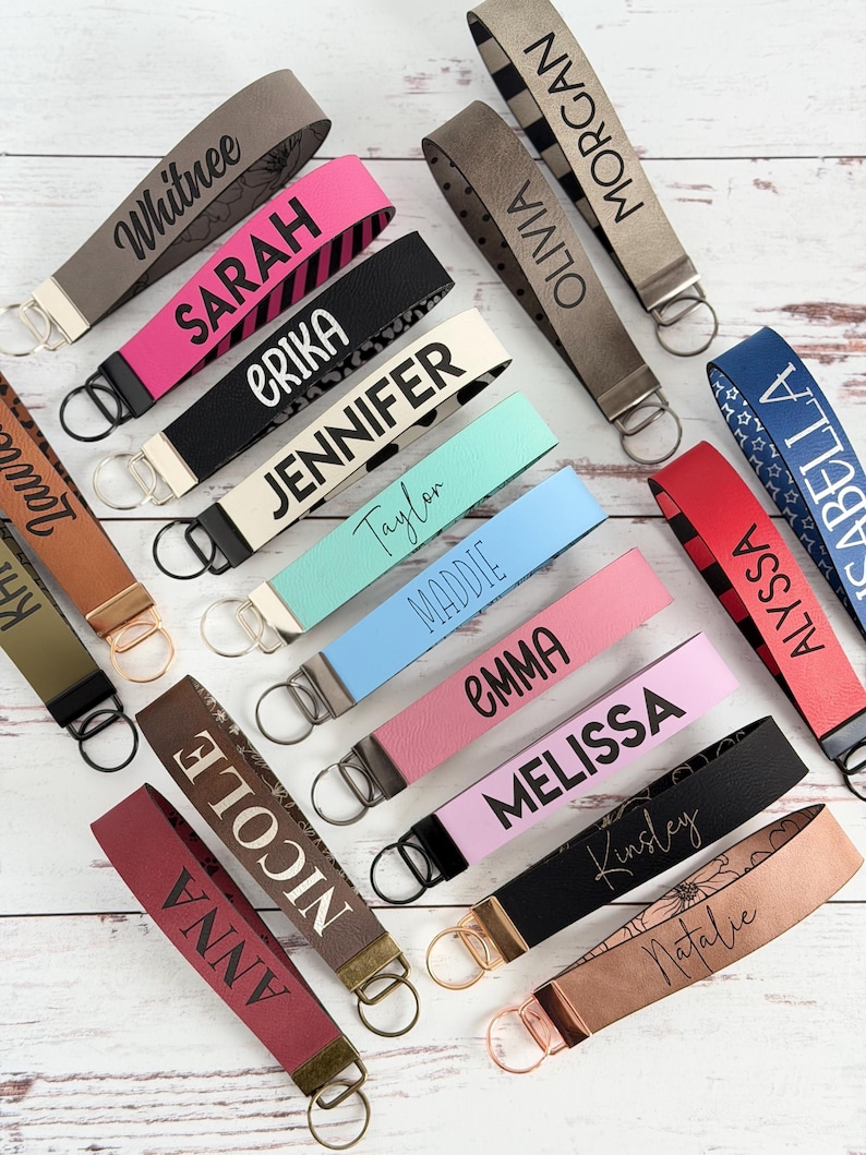 May include: A collection of personalized keychains in various colors, including pink, black, and blue. Each keychain features a name in a contrasting color, such as "Sarah" or "Jennifer", and includes a metal ring for attaching keys. The keychains are arranged on a light wood surface.