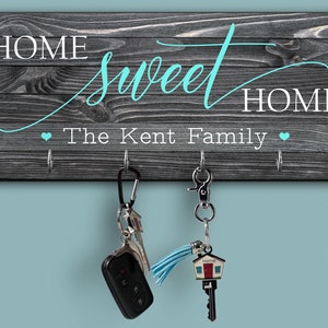 May include: A wooden key holder with the text "Home Sweet Home The Kent Family" in white lettering with a turquoise heart on either side of the text. There are four hooks on the holder with two keys hanging from them.
