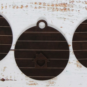 May include: Three round wooden ornaments with a brown woodgrain finish. Each ornament has a small house cutout with a heart in the center. The houses are painted white with a brown heart.