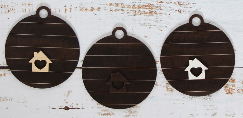 May include: Three round wooden ornaments with a brown woodgrain finish. Each ornament has a small house cutout with a heart in the center. The houses are painted white with a brown heart.