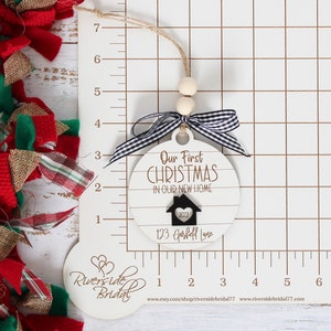 May include: White wooden Christmas ornament with black and white checkered ribbon. The ornament reads "Our First Christmas in Our New Home" with a house icon and the year 2022. The address "123 Garfield Lane" is written below the house icon.
