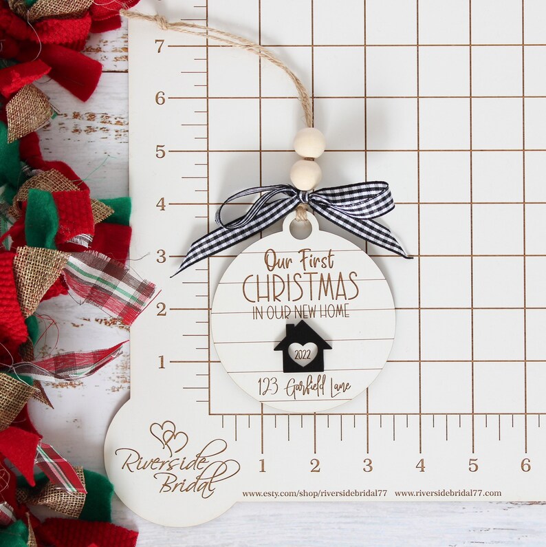 May include: White wooden Christmas ornament with black and white checkered ribbon. The ornament reads "Our First Christmas in Our New Home" with a house icon and the year 2022. The address "123 Garfield Lane" is written below the house icon.