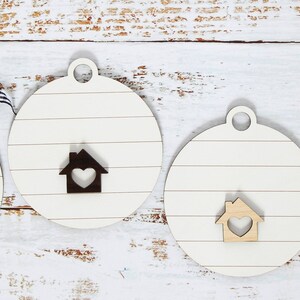 May include: Four round wooden ornaments with a whitewash finish and a house cutout with a heart in the center. One ornament has the text "Our First Christmas in Our New Home 2022 123 Garfield Lane" in black.