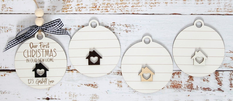 May include: Four round wooden ornaments with a whitewash finish and a house cutout with a heart in the center. One ornament has the text "Our First Christmas in Our New Home 2022 123 Garfield Lane" in black.