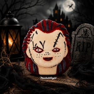 May include: A round bath bomb shaped like a doll head with pale skin, red hair, and dark stitched facial details. The surface features black painted scars, freckles, and red-rimmed eyes. The text Minibubblingbb is printed at the bottom.