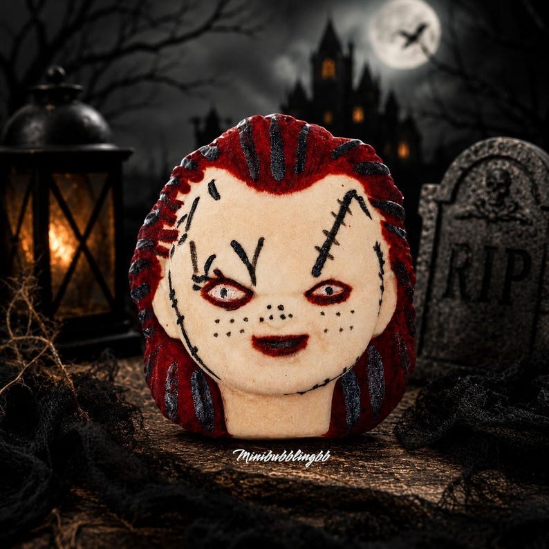 May include: A round bath bomb shaped like a doll head with pale skin, red hair, and dark stitched facial details. The surface features black painted scars, freckles, and red-rimmed eyes. The text Minibubblingbb is printed at the bottom.