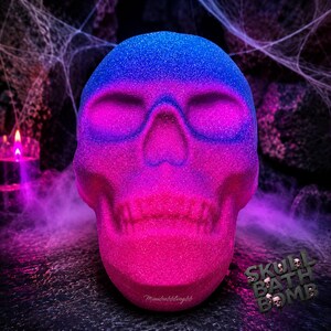 May include: A skull-shaped bath bomb with a glittery finish, featuring a vibrant gradient of blue at the top, transitioning into bright pink at the jaw. The surface has a textured, sparkling appearance. The text "SKULL BATH BOMB" is visible.