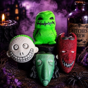 May include: A collection of four character-themed bath bombs including a round white face with a wide stitched grin, a bright green figure with a stitched mouth, a long green face with dark hair, and a red pointed face with a sharp-toothed grin.