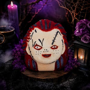 May include: A bath bomb shaped like a doll head with pale skin, red hair, and dark blue streaks. The face features painted black stitches, freckles, red lips, and red-rimmed eyes. The surface has a textured appearance with visible glittery accents.