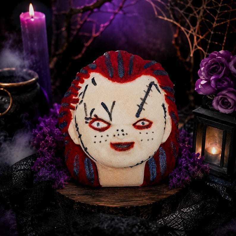 May include: A bath bomb shaped like a doll head with pale skin, red hair, and dark blue streaks. The face features painted black stitches, freckles, red lips, and red-rimmed eyes. The surface has a textured appearance with visible glittery accents.