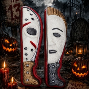 May include: Two vertical knife-shaped cookies. The left cookie features a white hockey mask with black holes and red accents, and a tan handle. The right cookie features a white human face mask with brown hair and a dark grey textured handle.