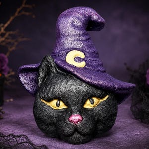 May include: A black cat head figurine wearing a purple pointed witch hat with a yellow crescent moon emblem. The cat has textured black fur, bright yellow almond-shaped eyes with defined eyeliner, and a small pink nose, crafted in a solid form.