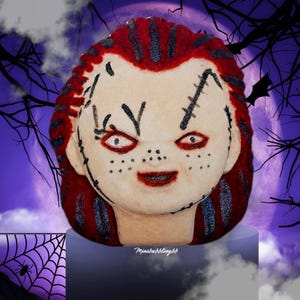 May include: A round, pale-skinned doll head bath bomb with bright red hair featuring dark glittery streaks. The face has painted black stitches, dark red eye makeup, and a small red mouth. The text Mimibubblingbb is printed at the base.