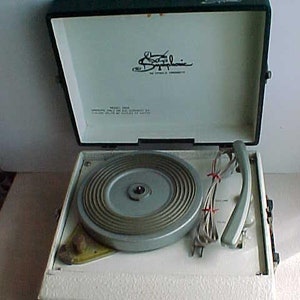 May include: Vintage portable record player with a white textured exterior and a dark green lid. The turntable is silver, and the lid interior features a logo and text. The record player has a handle and various cords.