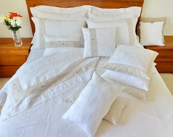 May include: White linen bedding with embroidered details. The bedding includes a duvet cover, pillowcases, and decorative throw pillows. The pillows have a beige trim and are arranged on a white bed.