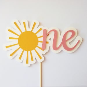 May include: A daisy-shaped cake topper with a yellow center and white petals. The word "one" is written in pink script. The topper is attached to a wooden stick, perfect for a first birthday celebration.