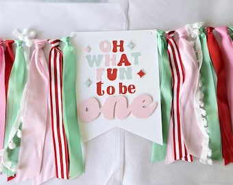 May include: A festive banner with the words "Oh What Fun to be One" in pink, red, and green letters. The banner is decorated with red, pink, and green ribbon streamers.