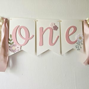 May include: A banner with the word "one" in pink script letters on white pennants. The banner is decorated with floral accents and colorful ribbons with pom-poms, perfect for a first birthday celebration.