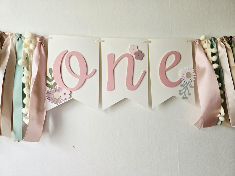 May include: A banner with the word "one" in pink script letters on white pennants. The banner is decorated with floral accents and colorful ribbons with pom-poms, perfect for a first birthday celebration.