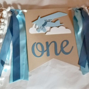 Puede incluir: Una pancarta azul y marr&oacute;n con pompones blancos y cintas azules y blancas. La pancarta dice "one" en una fuente blanca de escritura.