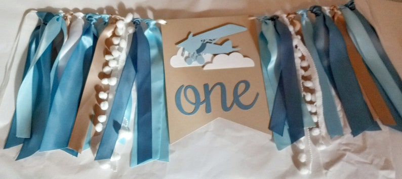 Puede incluir: Una pancarta azul y marr&oacute;n con pompones blancos y cintas azules y blancas. La pancarta dice "one" en una fuente blanca de escritura.