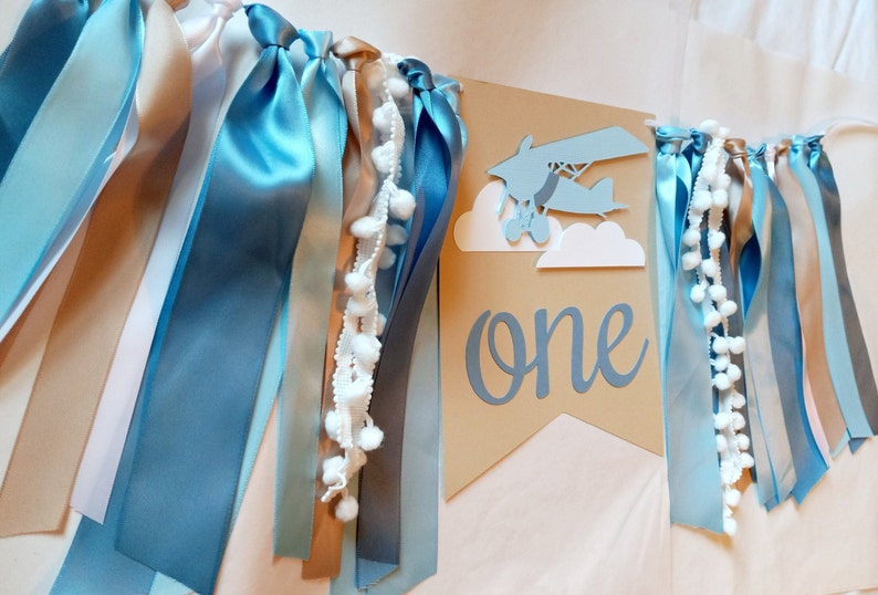 Puede incluir: Una pancarta de papel marr&oacute;n con la palabra "one" en letra azul. Un avi&oacute;n azul con nubes blancas est&aacute; por encima de la palabra. La pancarta est&aacute; decorada con cintas azules, blancas y marrones.