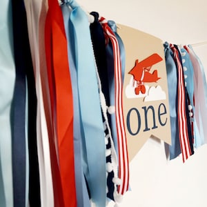 K&ouml;nnte beinhalten: Ein braunes Banner mit dem Wort "one" in wei&szlig;en Buchstaben und einem roten Flugzeug mit wei&szlig;en Wolken. Das Banner ist mit blauen, roten und wei&szlig;en B&auml;ndern verziert.