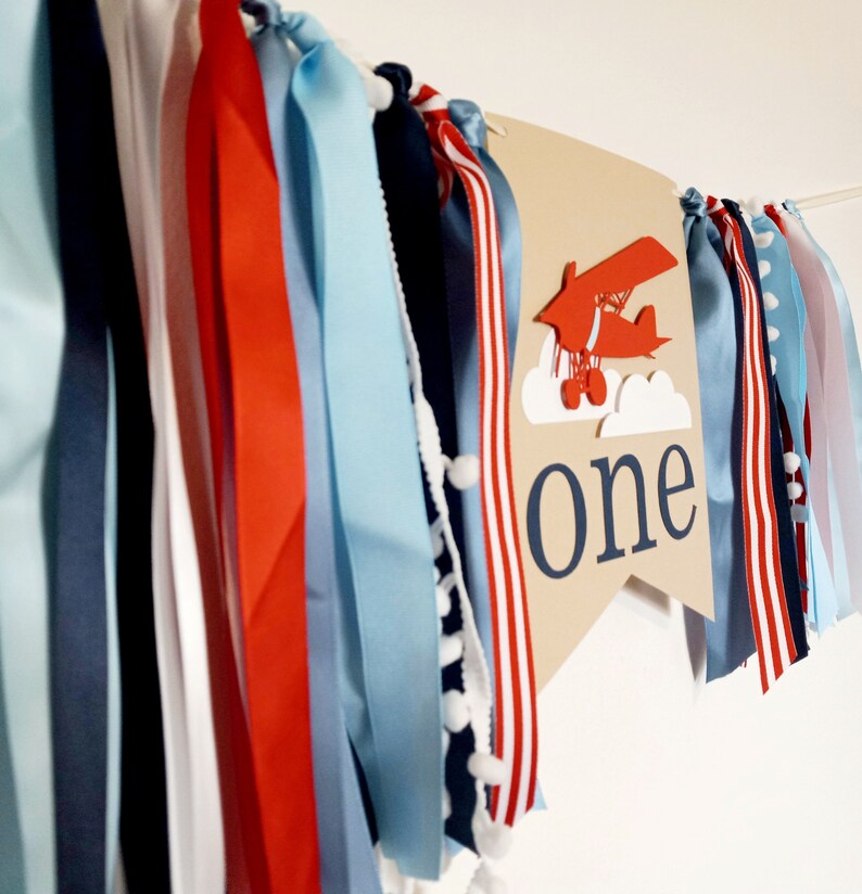 K&ouml;nnte beinhalten: Ein braunes Banner mit dem Wort "one" in wei&szlig;en Buchstaben und einem roten Flugzeug mit wei&szlig;en Wolken. Das Banner ist mit blauen, roten und wei&szlig;en B&auml;ndern verziert.