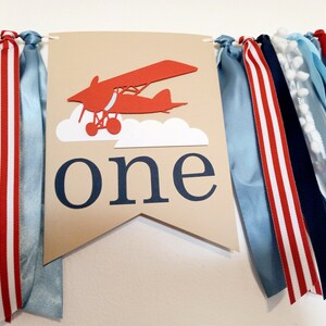 K&ouml;nnte beinhalten: Ein braunes Banner mit einem roten Flugzeug und wei&szlig;en Wolken, das Wort "eins" in blauen Buchstaben und eine Girlande aus blauen, wei&szlig;en und roten B&auml;ndern.
